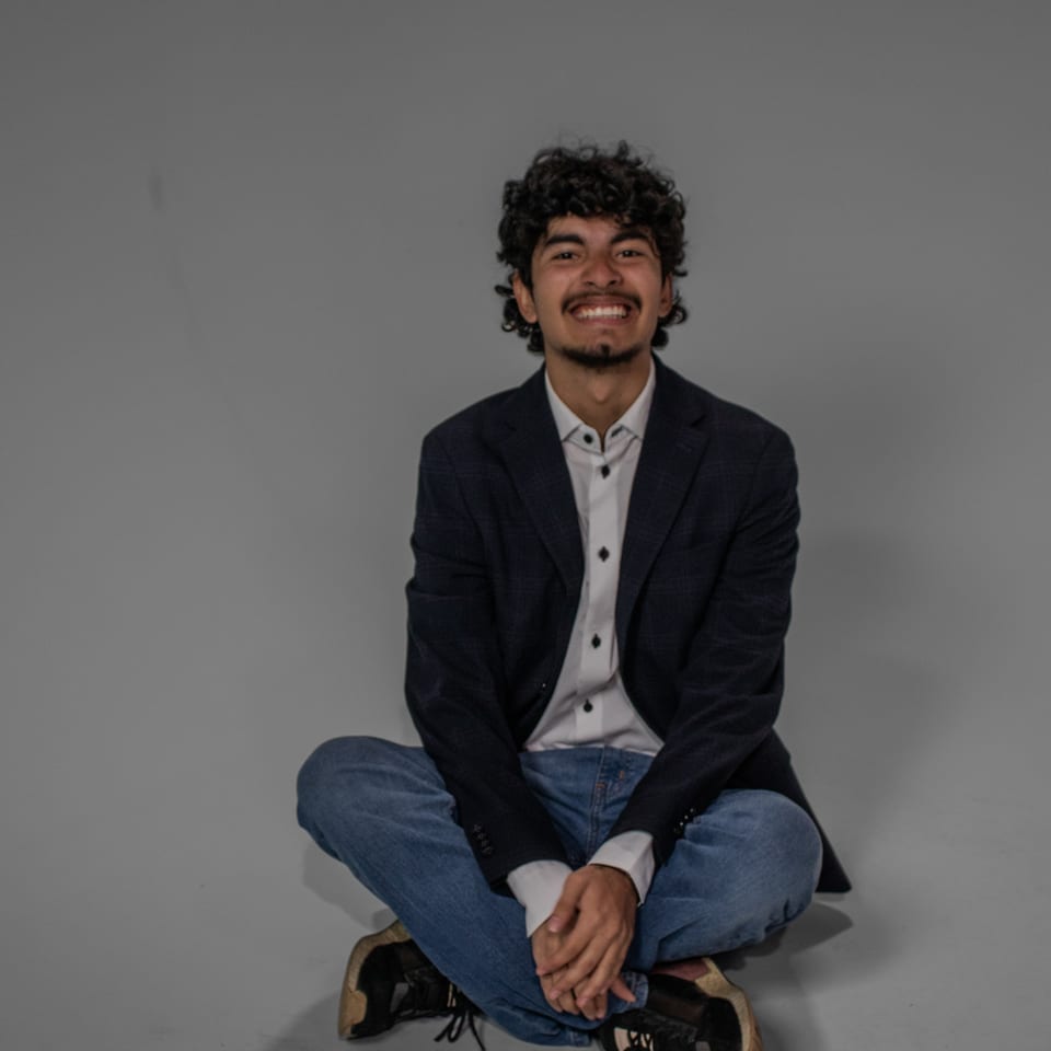 Young man in a navy blazer and jeans sitting cross-legged on the studio floor, grinning.
