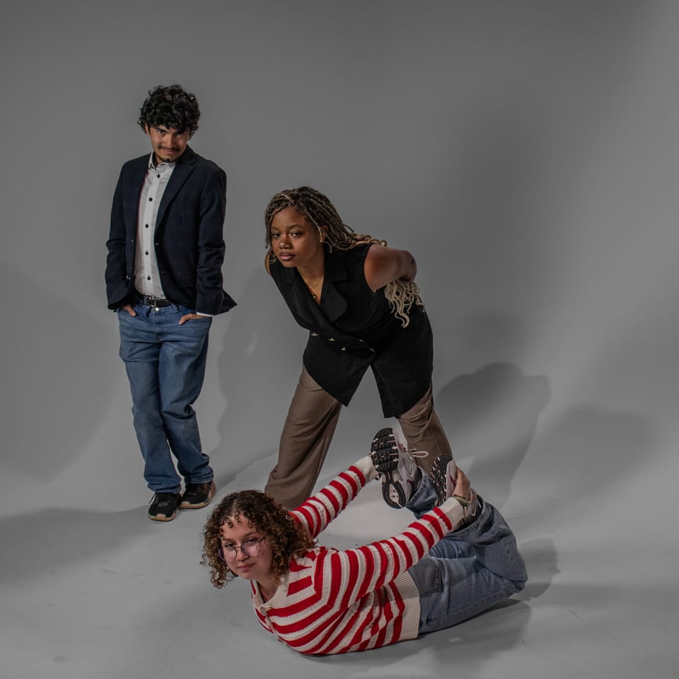 Three friends clowning around: one standing with his hands in his pockets, one bent over laughing, one lying on the floor with her feet in the air.