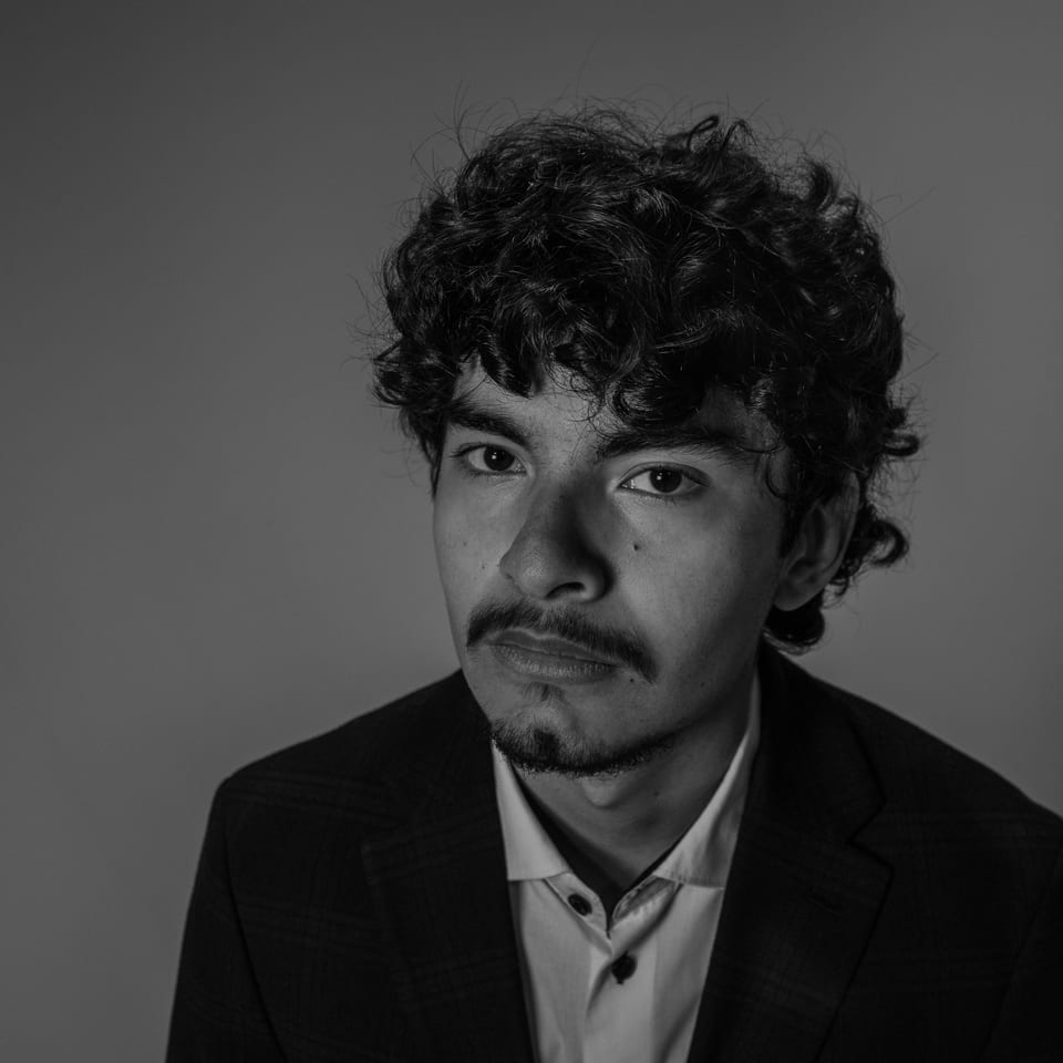 Black-and-white headshot of a young man with curly hair and a moustache, giving the camera a skeptical look.