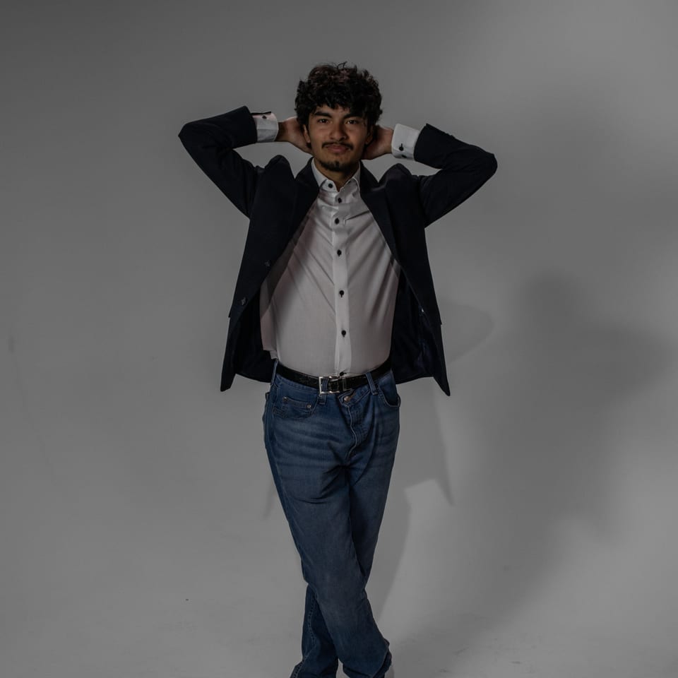 Full-length portrait of a young man in a blazer and jeans, hands behind his head, legs crossed.
