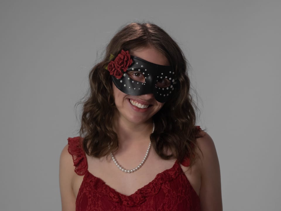 Woman in a black masquerade mask with a red rose, smiling in a red dress and pearls.