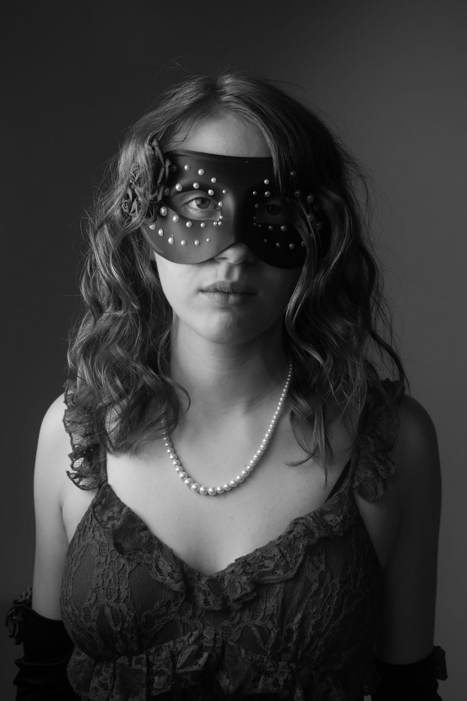 Black-and-white portrait of a woman in a studded masquerade mask and a pearl necklace.