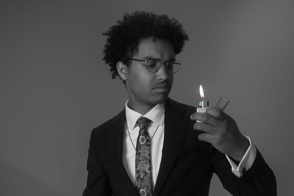 Black-and-white portrait of a man in a suit and patterned tie, studying the flame of a lighter.