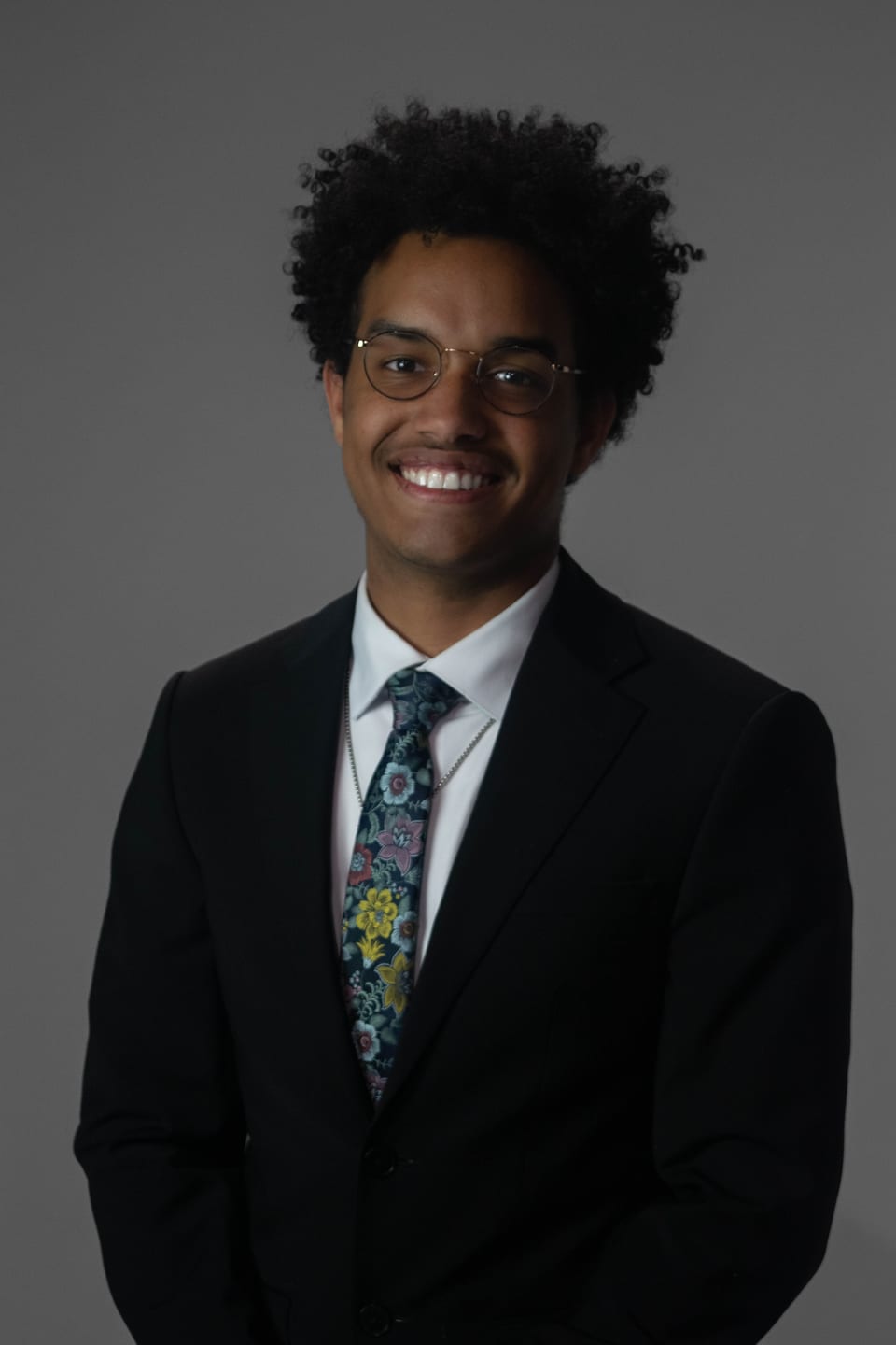 Smiling young man in glasses, a dark suit and a floral tie.