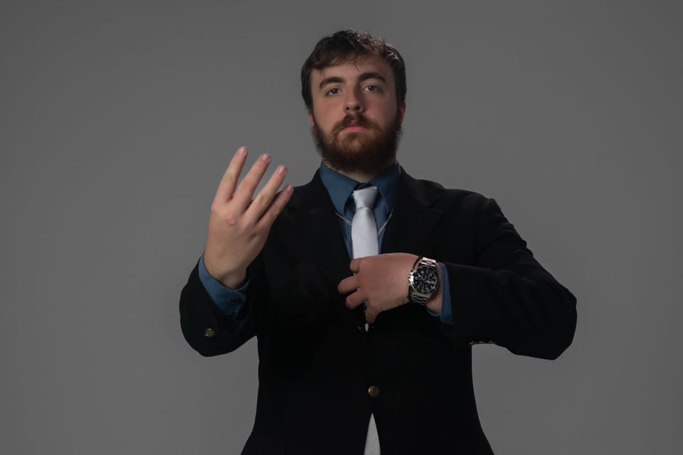 Bearded man in a dark blazer raising one open hand toward the camera, a watch on his wrist.