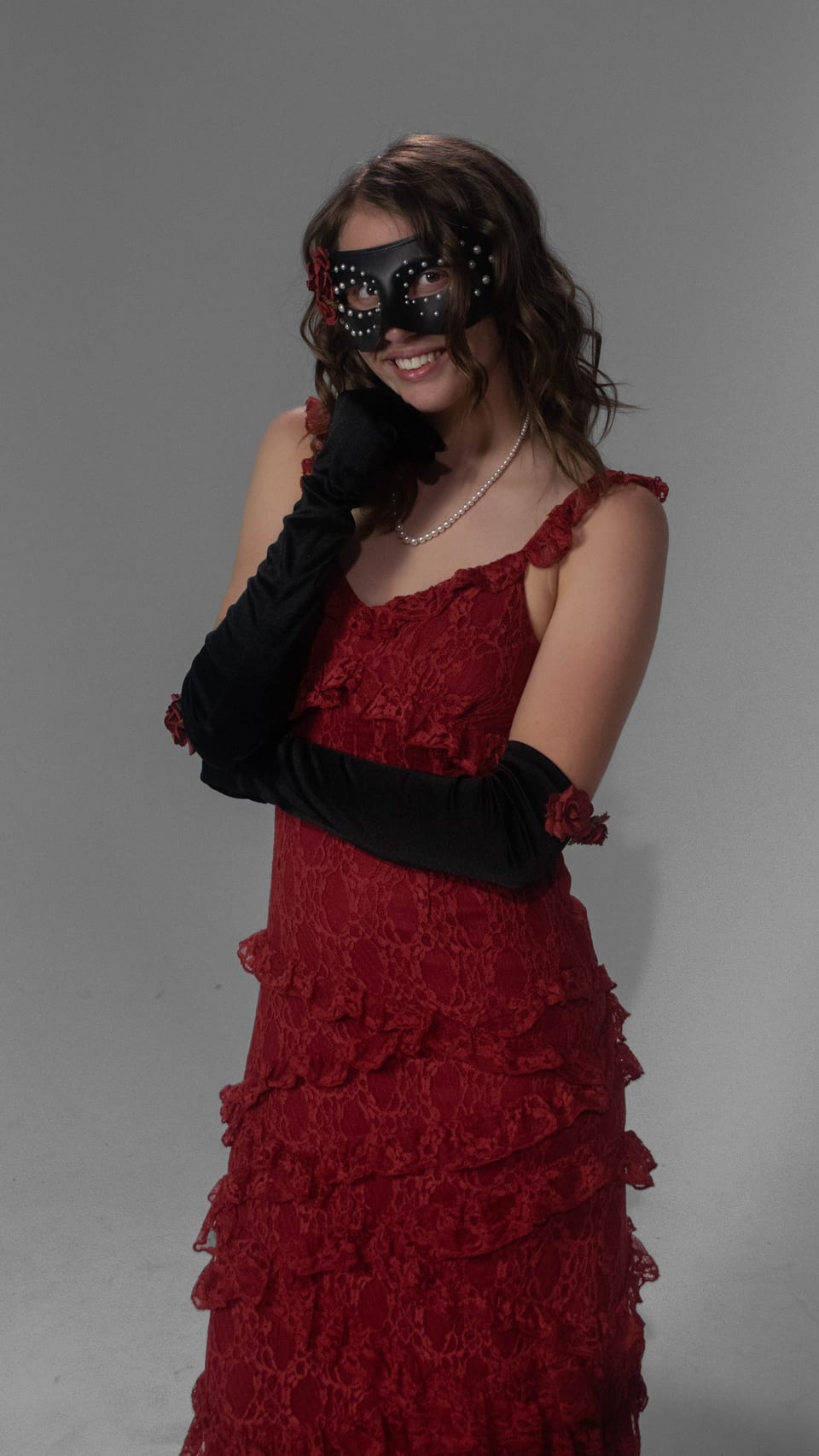 Woman in a red ruffled gown, masquerade mask and long black gloves, one hand resting against her cheek.