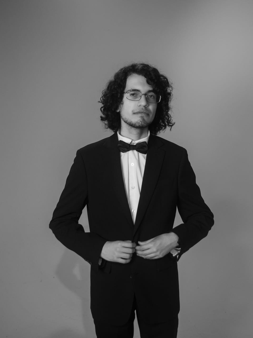 Black-and-white full-length portrait of a young man with long curly hair in a tuxedo and bow tie, buttoning his jacket.