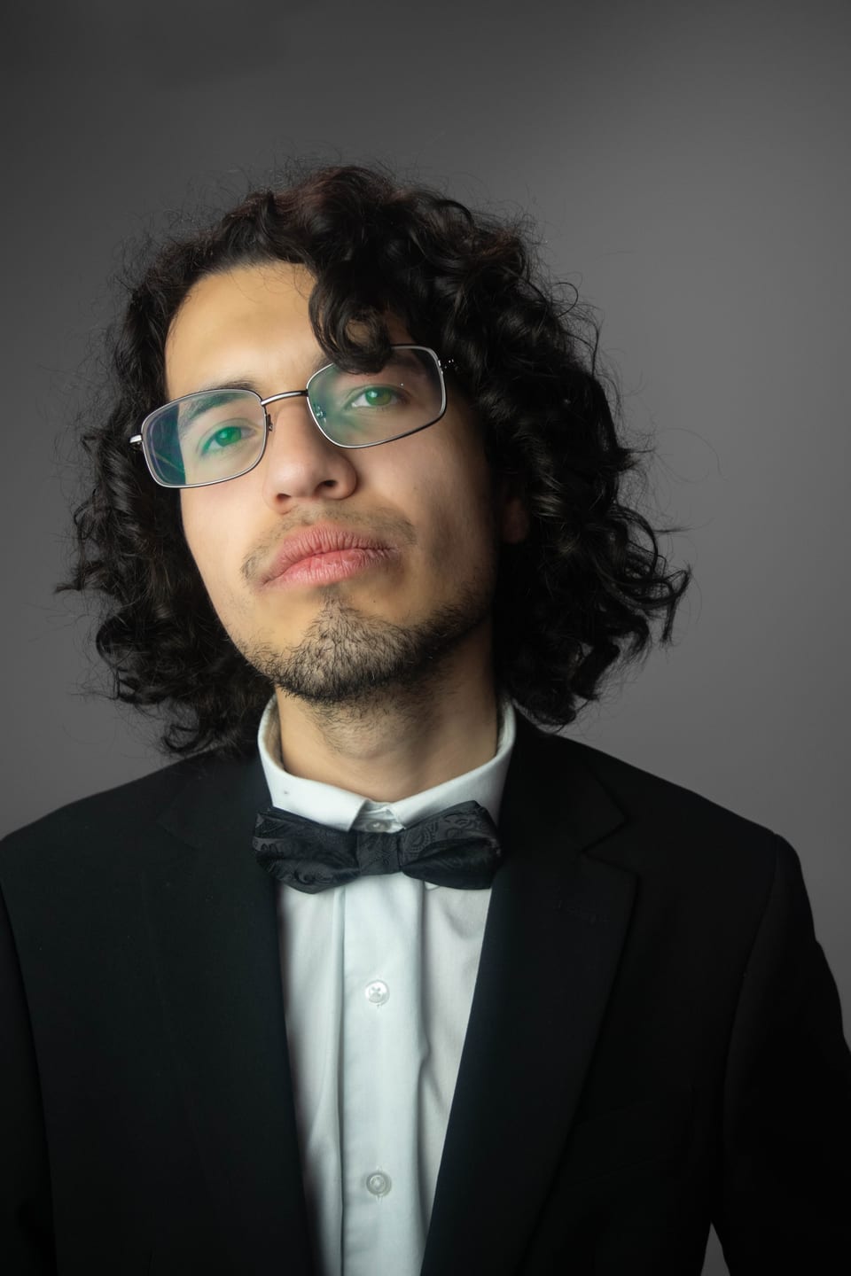 Close portrait of a man with long curly hair, wire-rimmed glasses and a bow tie.