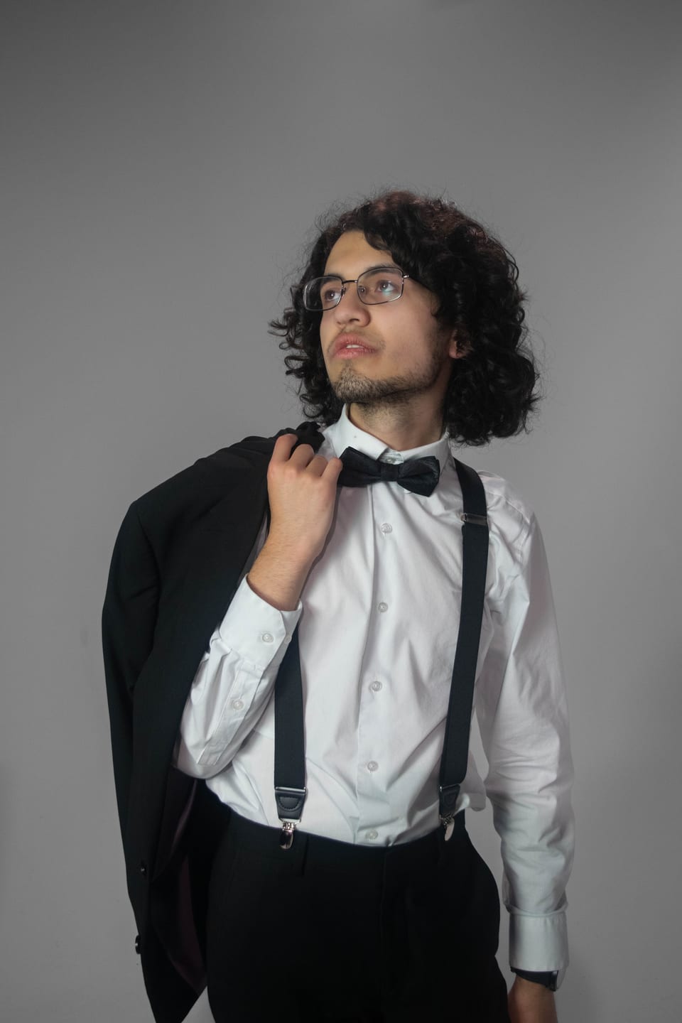 Young man in suspenders and a bow tie with his jacket over his shoulder, looking up and away.