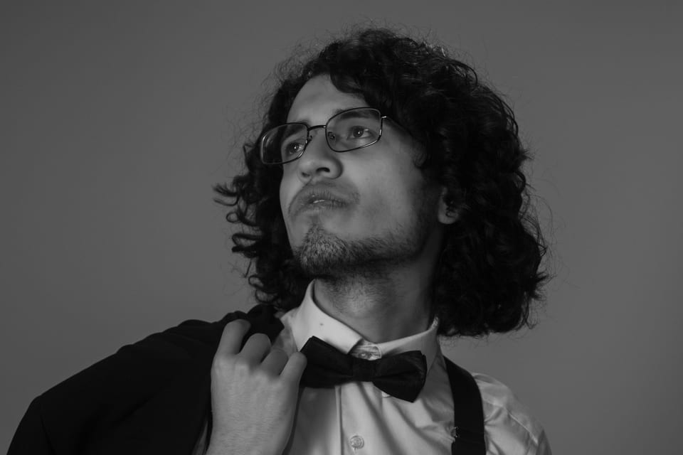 Black-and-white portrait of a man with long curly hair and glasses looking upward, jacket over his shoulder.