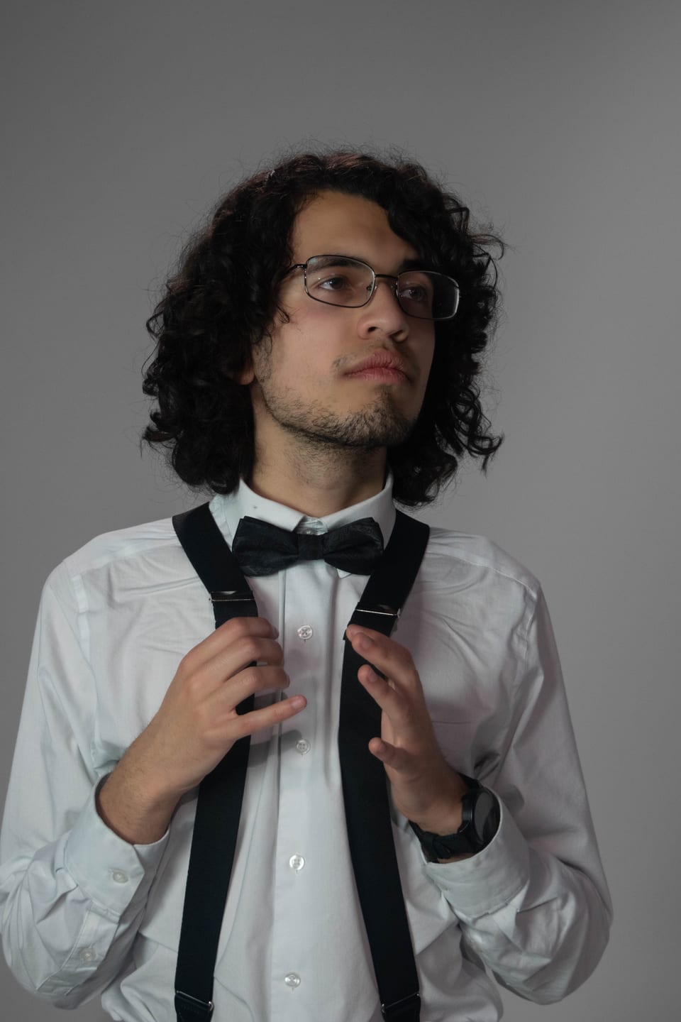 Young man with long curly hair and glasses adjusting his suspenders.