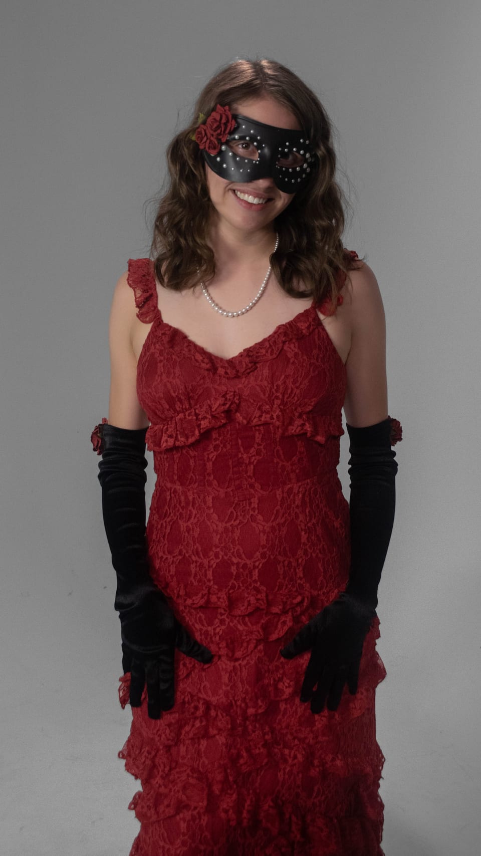 Full-length portrait of a woman in a red ruffled gown, long black gloves and a masquerade mask.