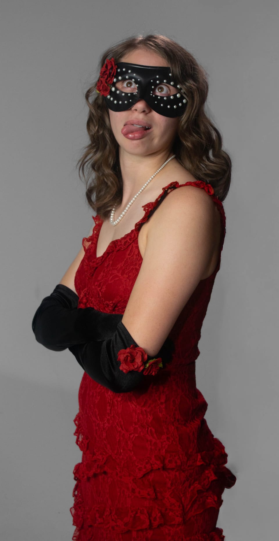 Woman in a red gown and masquerade mask sticking her tongue out, arms crossed.