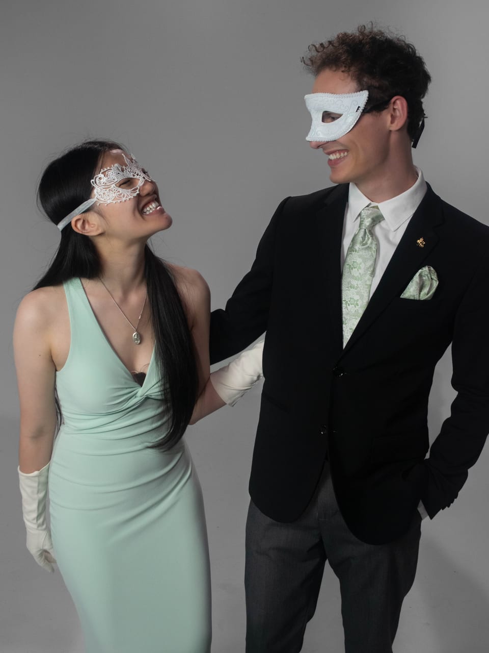 A couple in masquerade masks laughing at each other; she wears a mint gown and white gloves, he wears a dark suit.