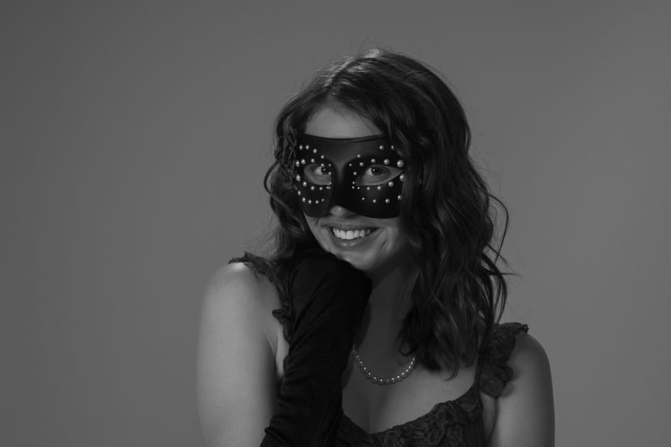 Black-and-white photo of a smiling woman in a studded mask, her gloved hand under her chin.