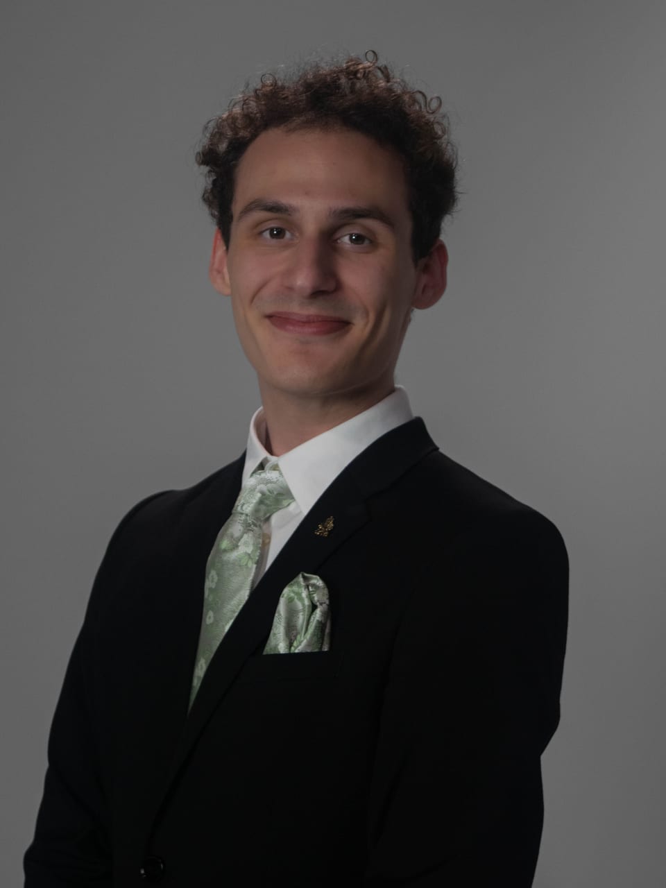Smiling young man with curly hair in a dark suit and sage-green tie.
