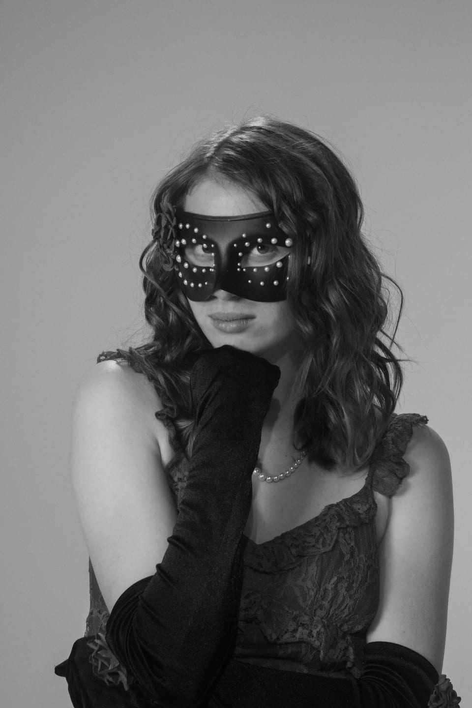 Black-and-white portrait of a woman in a studded mask resting her chin on a long black glove.