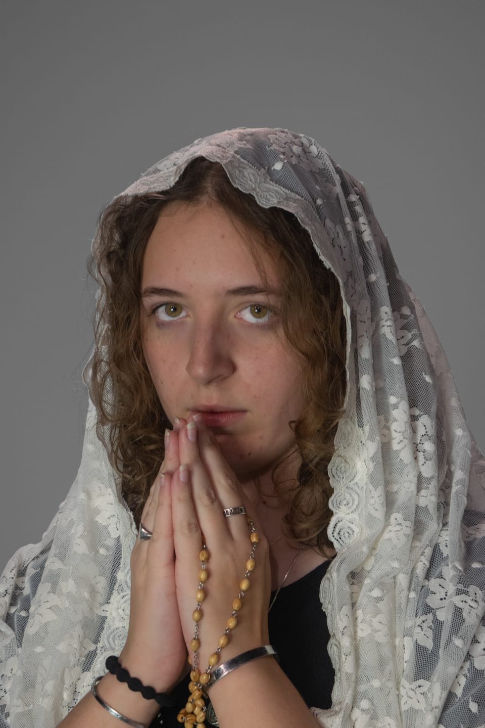 Woman in a white lace veil with her hands pressed together in prayer, a wooden rosary between her fingers.