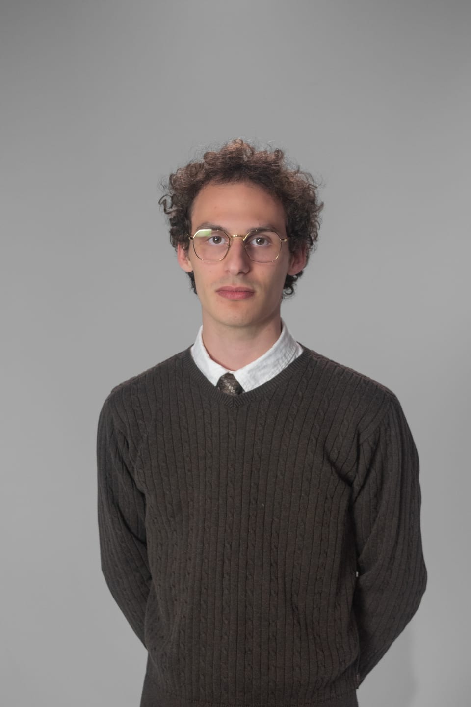 Young man in glasses, a knit sweater and a collared shirt standing with his arms at his sides.