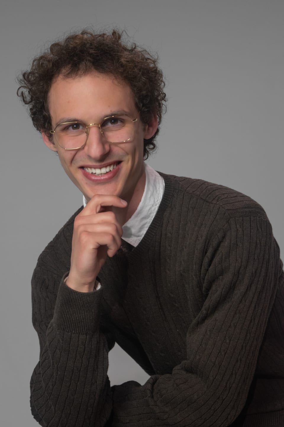 Grinning young man in glasses and a knit sweater, chin resting on his hand.