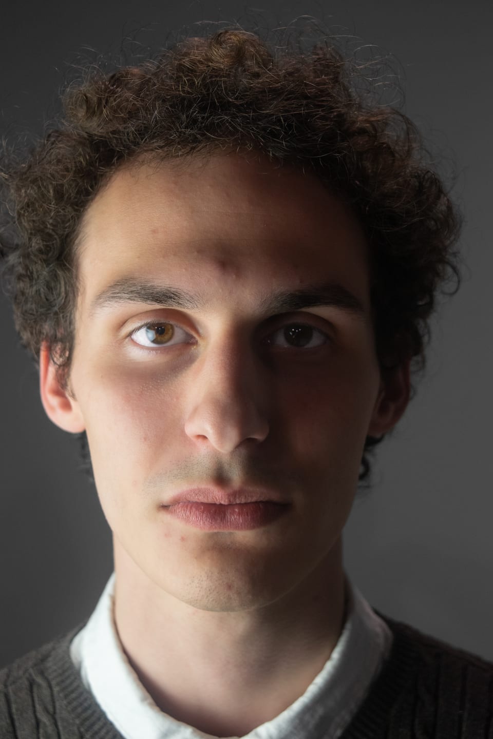 Tight portrait of a young man with curly hair, lit from one side against a dark backdrop.