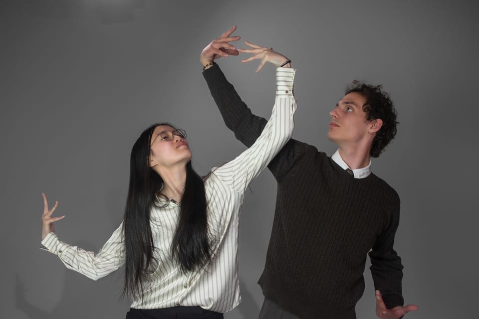 Two dancers mid-pose, arms stretched overhead, reaching past each other.