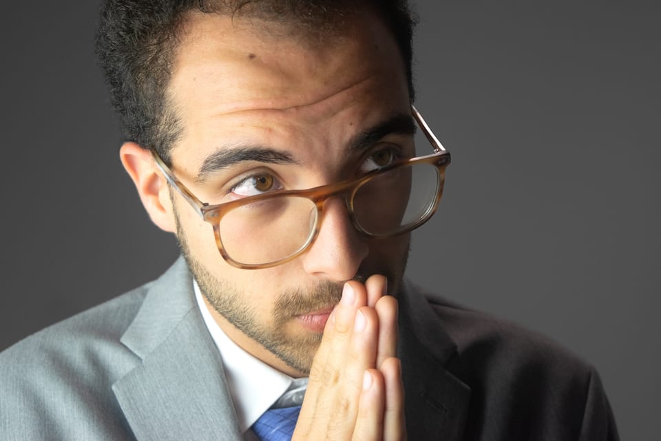 Man in tortoiseshell glasses and a grey suit, hands pressed together at his lips, eyes turned upward.