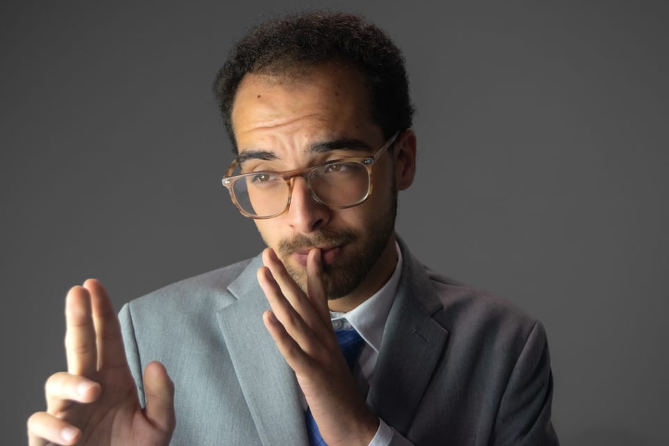 Man in tortoiseshell glasses and a grey suit, one hand at his lips and the other raised with two fingers out.