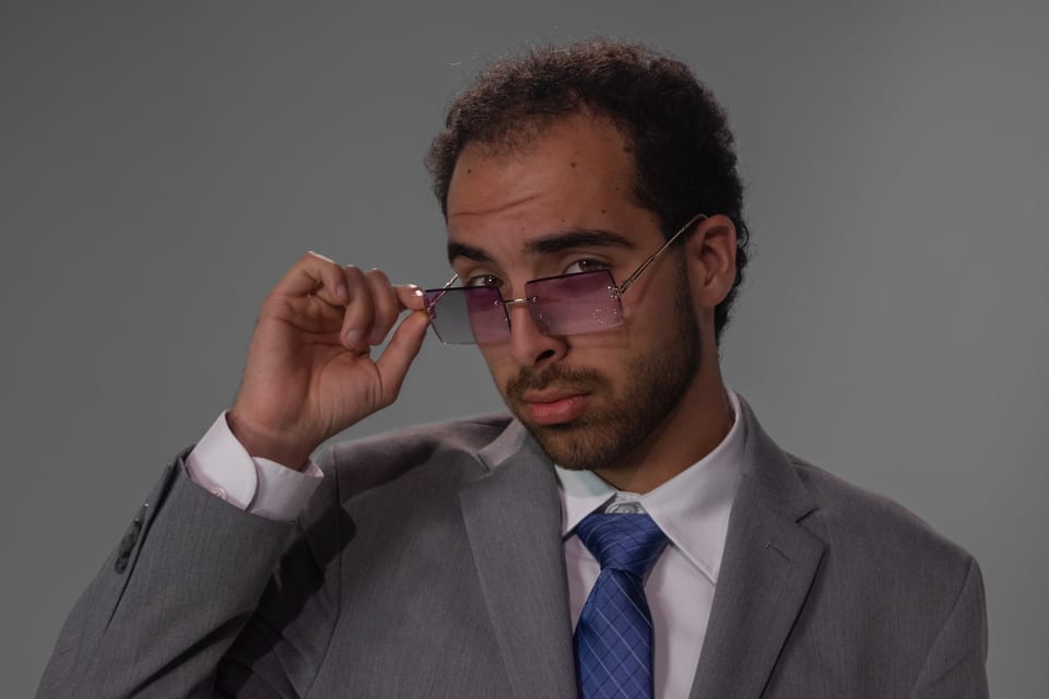 Man in a grey suit and blue tie peering over purple-tinted sunglasses.
