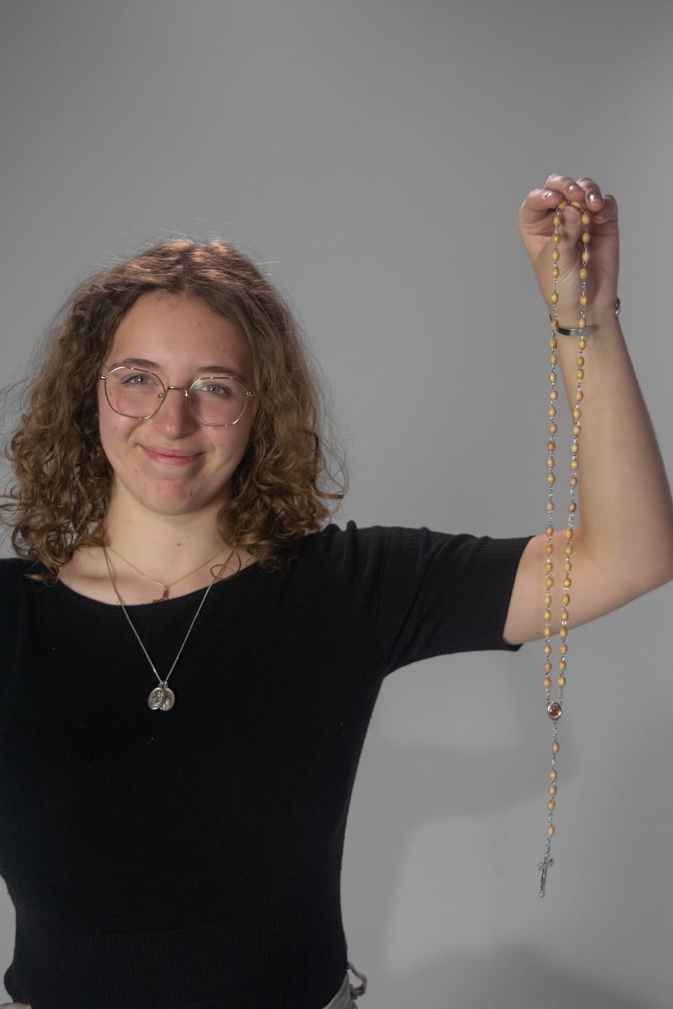 Smiling woman with curly hair holding a wooden rosary high in one hand.