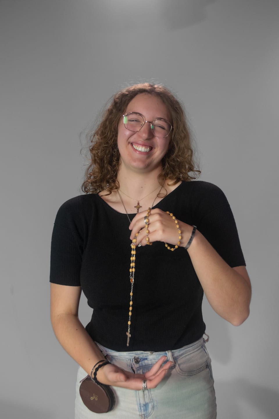 Beaming woman in glasses holding a wooden rosary against her chest.