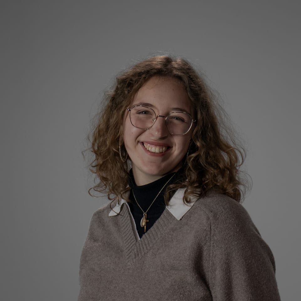 Smiling woman with curly hair and glasses in a sweater over a black turtleneck.