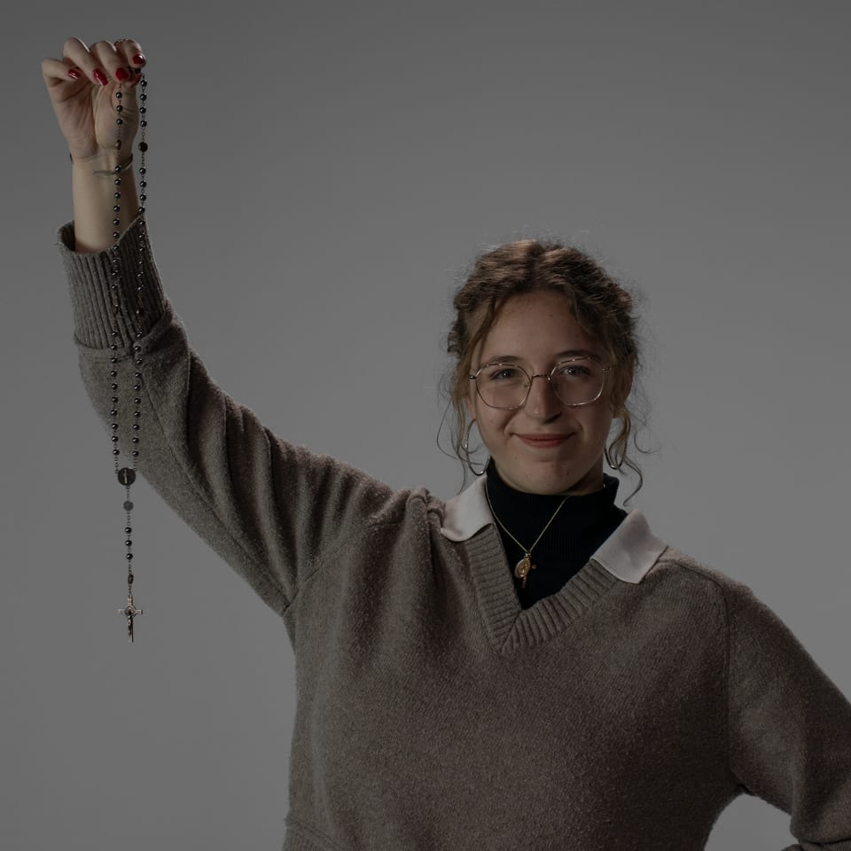 Smiling woman in glasses holding a dark rosary high above her head.