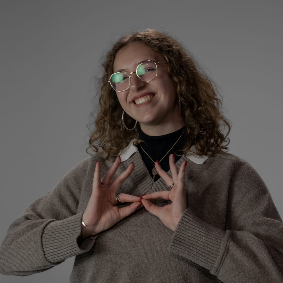 Smiling woman with curly hair and glasses making a heart shape with her hands.