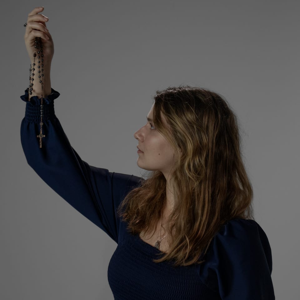 Woman in a navy dress seen in profile, holding a rosary aloft.