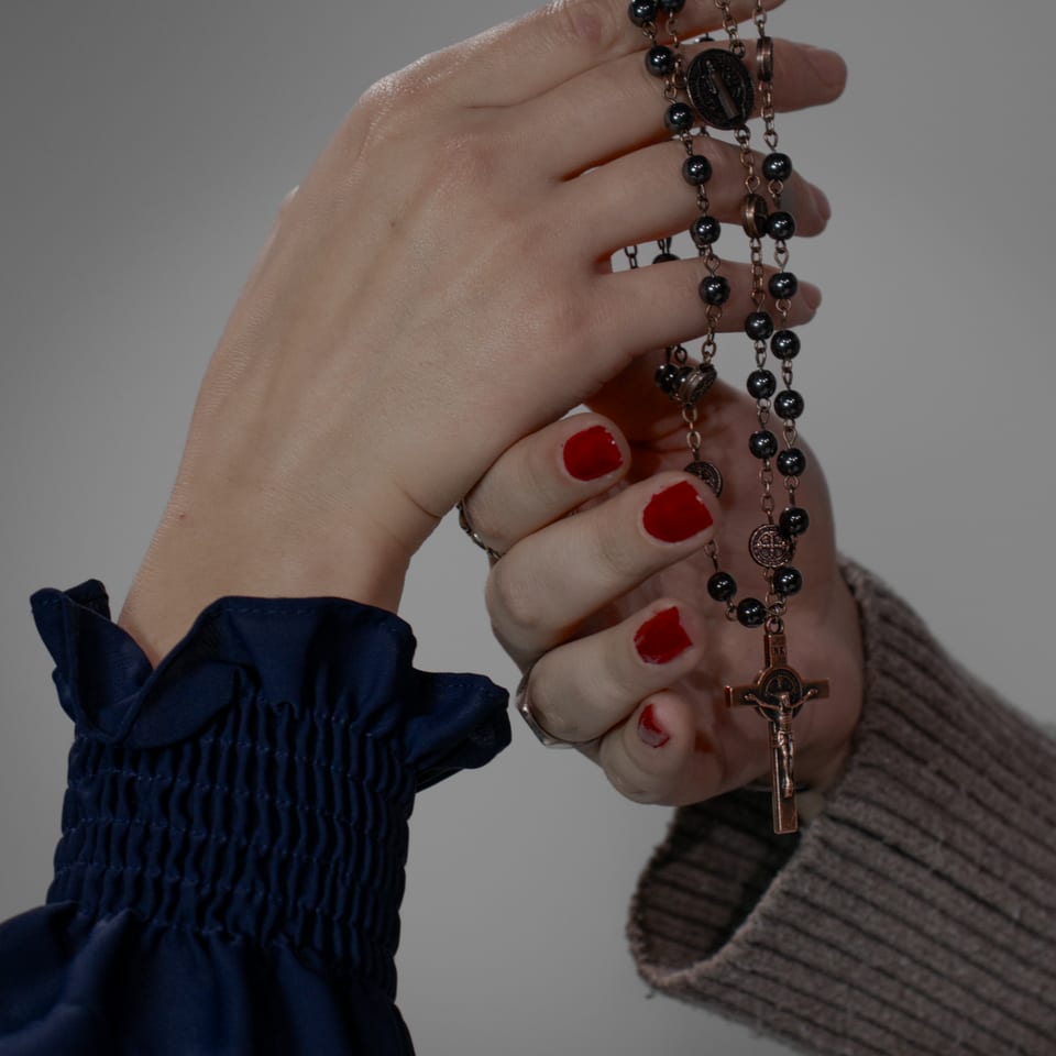 Close-up of two people's hands holding a dark rosary, one with red nail polish.
