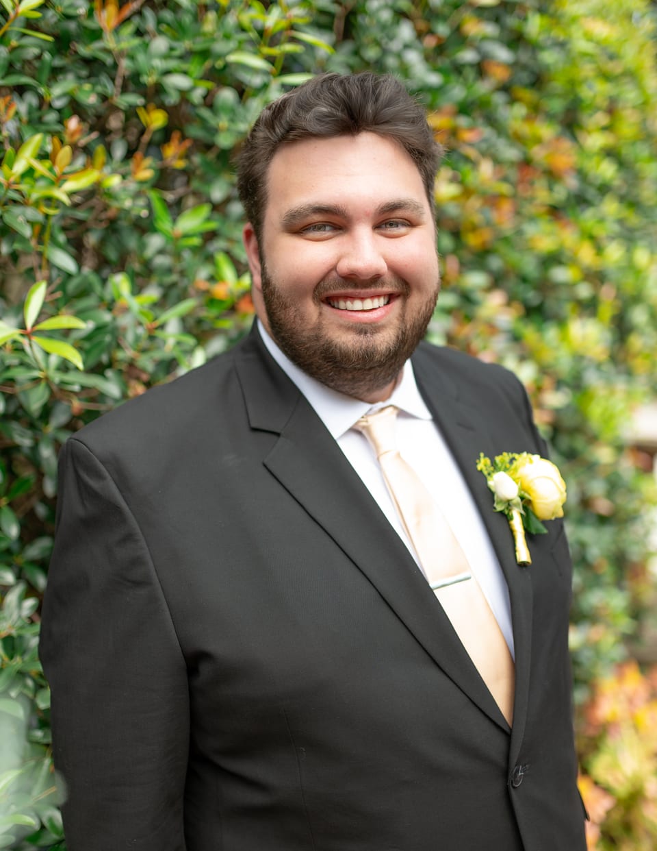 Ryan Bibby smiling in a dark suit with a yellow rose boutonnière, in front of a hedge.