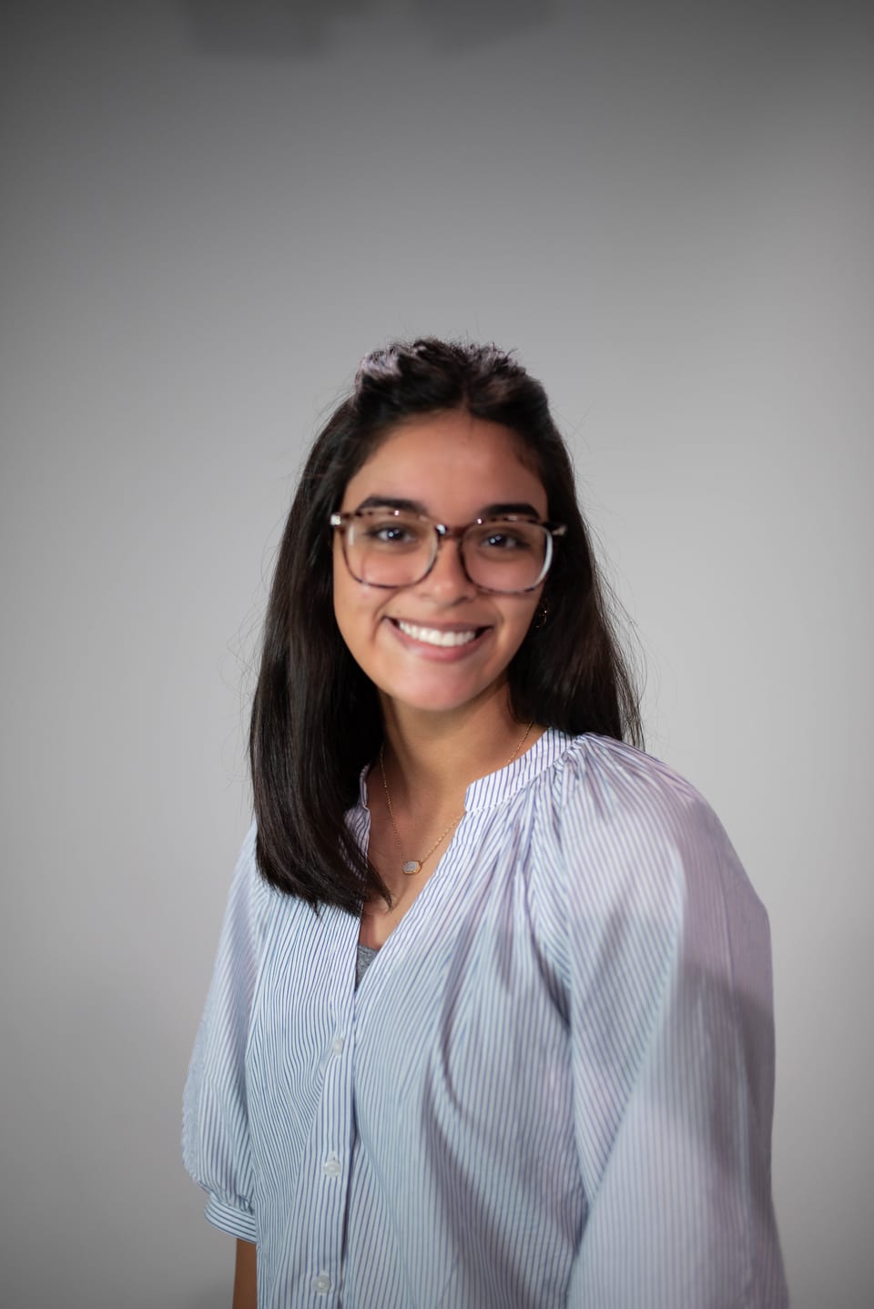 Smiling young woman with glasses and shoulder-length dark hair in a pale blue striped blouse.