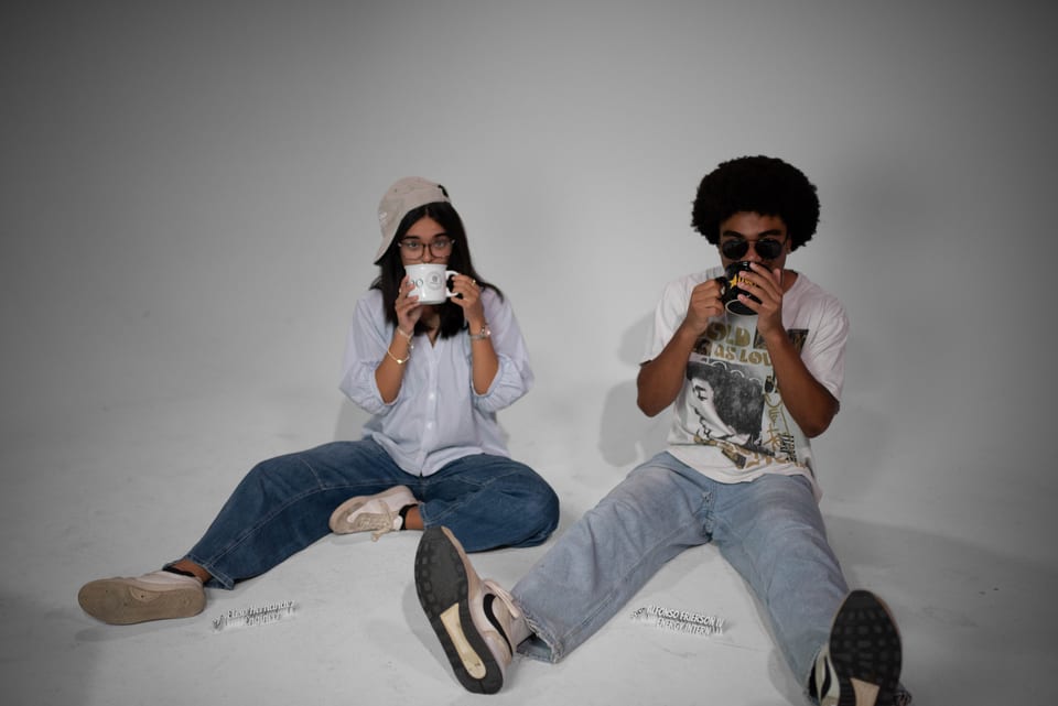 Two coworkers sitting on the studio floor sipping from mugs, one in a bucket hat, one in sunglasses.