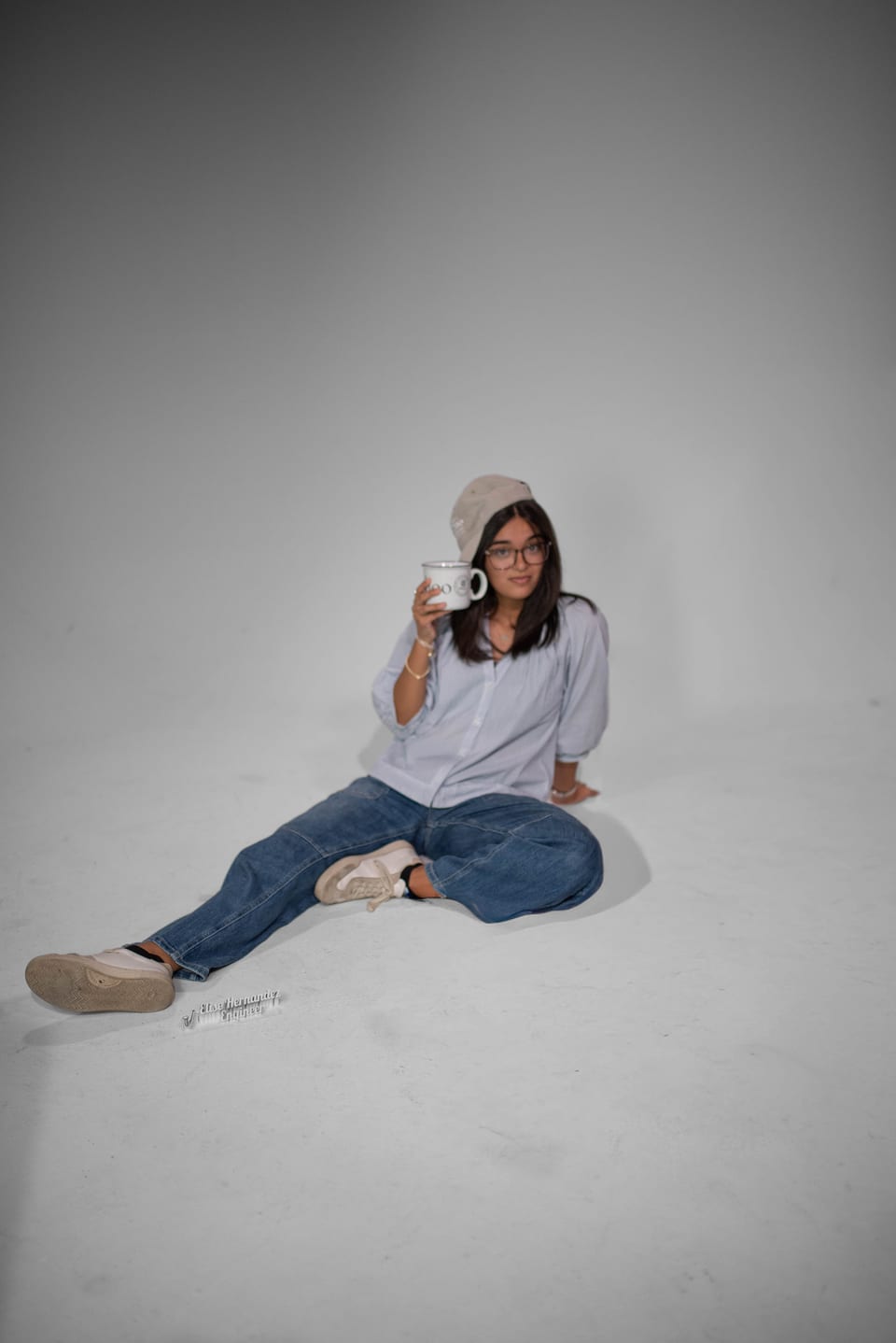 Young woman in a bucket hat sitting on the studio floor, sipping from a mug.