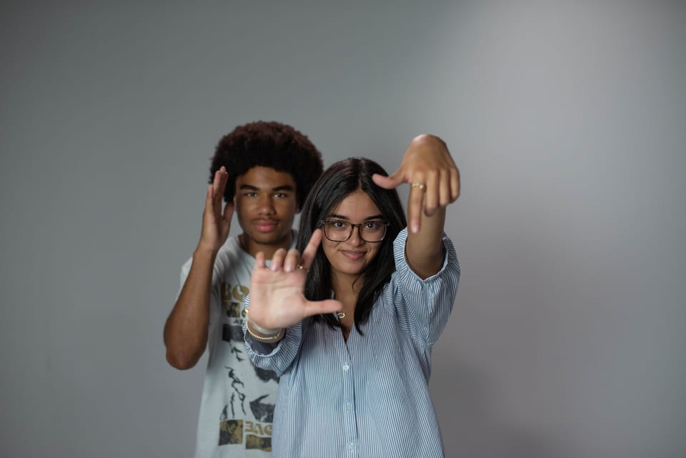 A young woman reaching an open hand toward the camera, a young man grinning over her shoulder.