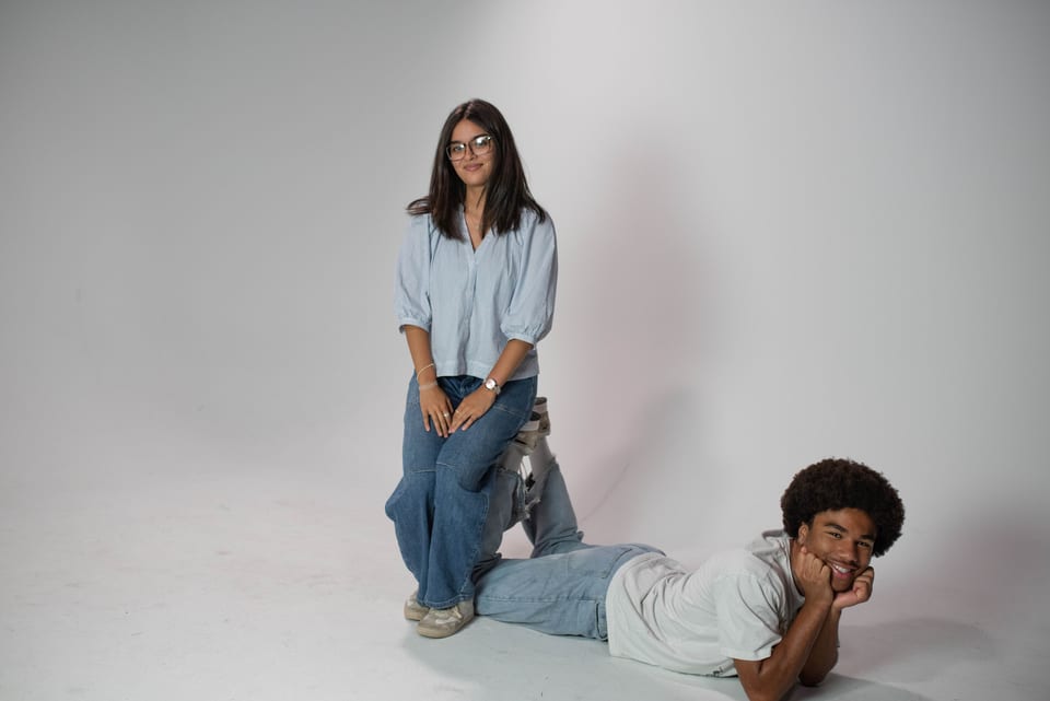 A young woman sitting on one knee while a young man lies on the floor behind her, chin in hand.
